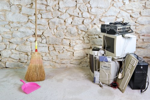 Inside a room with a stone wall background, there is a bundle of cleaning tools including a broom with a wooden handle and straw bristles, and a small pink dustpan, both positioned on the concrete floor in the foreground. To the right, a pile of electronic waste and household items, such as an old CRT television, computer tower, monitor, keyboard, and various cables, are stacked next to a small black speaker and a black rectangular box. The environment suggests a space prepared for rubbish removal or office clearance, typical of services provided by companies like Office Clearance Battersea based near Battersea, London. The lighting appears natural, illuminating the scene clearly, emphasizing the contrast between the clean, textured stone wall and the cluttered foreground objects. The overall setting reflects typical rubbish collection efforts in domestic or small commercial environments, aligning with local rubbish clearance services in Battersea or nearby areas of Wandsworth.