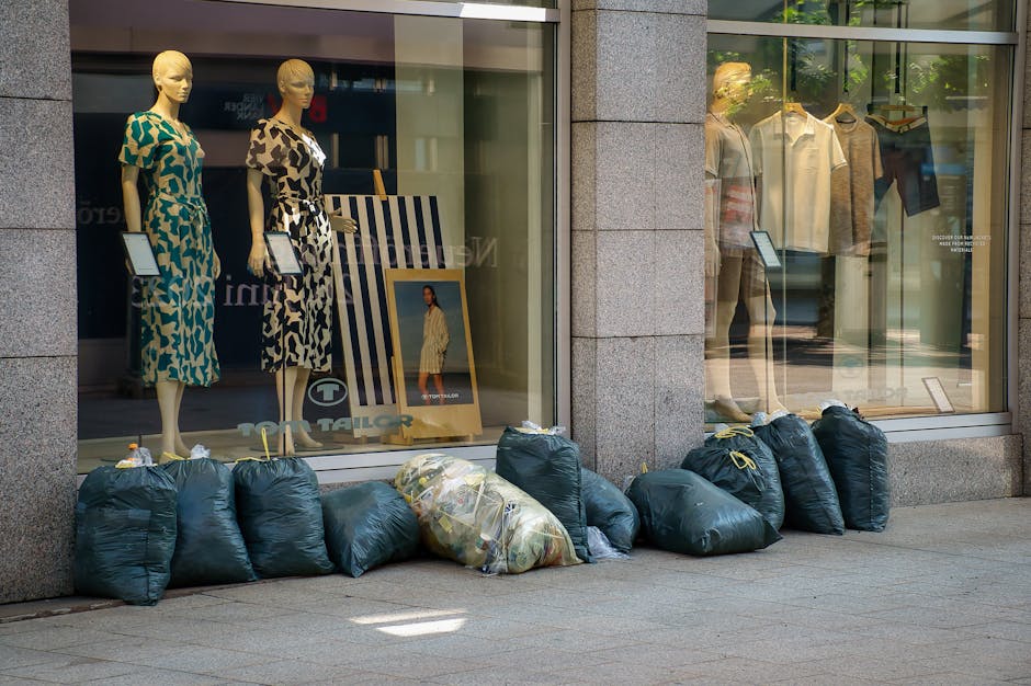 The image shows a row of three large wheeled rubbish bins positioned along the exterior wall of a building in an urban setting. The first bin on the left is gray with a closed lid, while the middle an