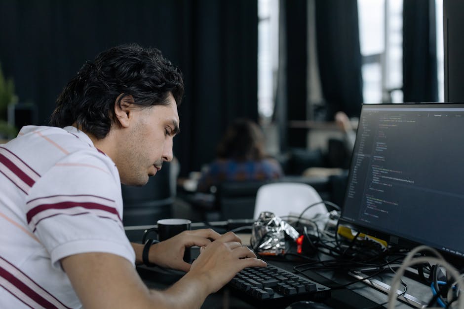 A close-up view of a silver laptop with a black keyboard, positioned on a light wooden desk. The laptop screen displays lines of programming code with syntax highlighting on a dark background. To the 