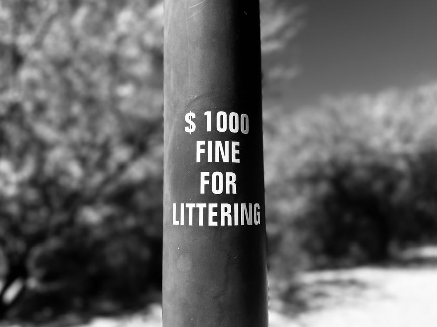 Close-up of a black cylindrical pole with a warning sign in white text stating a fine of $1000 for littering. The sign is centered on the pole, which appears to be made of metal or painted plastic, wi