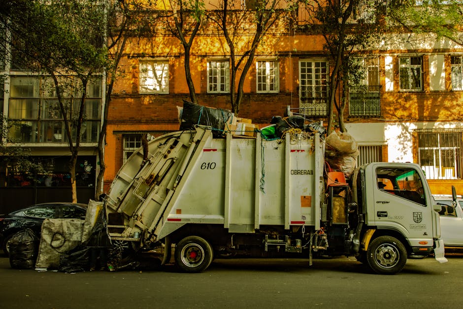A large outdoor area shows an overflowing collection of rubbish and waste materials placed on a paved surface. The assortment includes flattened cardboard boxes, plastic bags filled with mixed waste, 