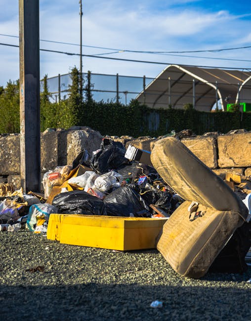 Two large black plastic bags filled with rubbish are placed on a pavement next to a black metal fence and dense foliage. One bag appears to contain paper or cardboard, with visible printed markings on